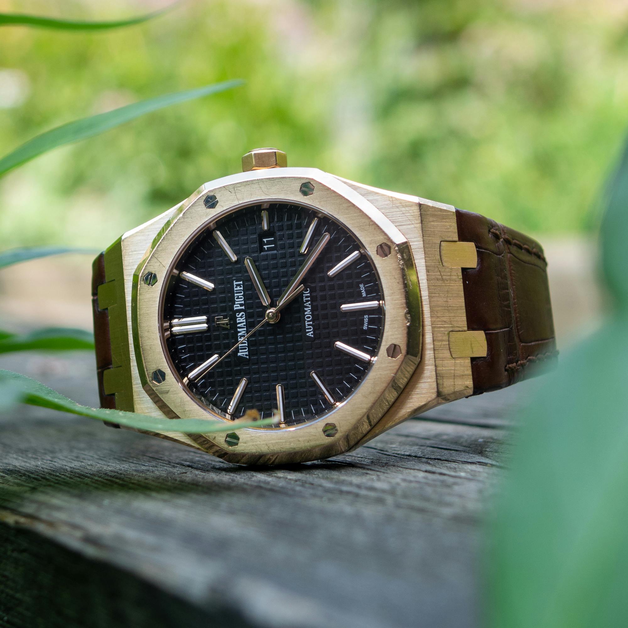 Audemars Piguet Royal Oak watch with a black dial and gold case on a wooden surface outdoors.