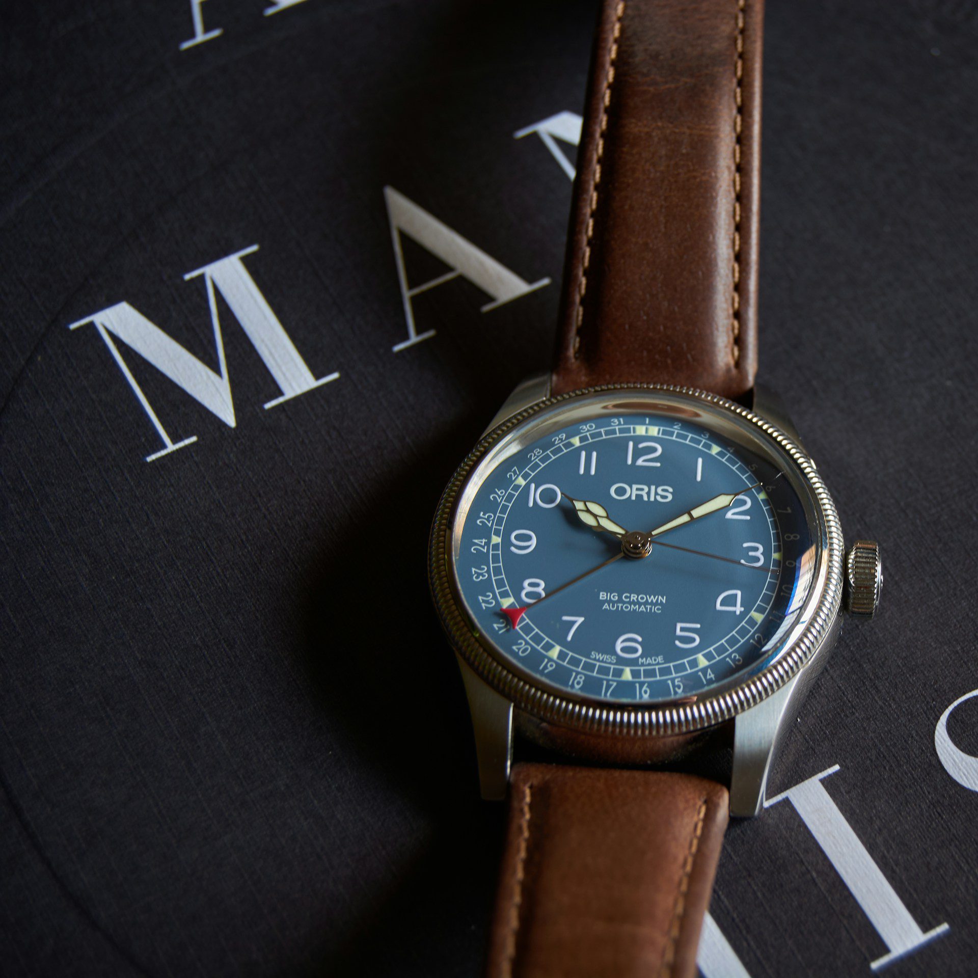 Close-up of an Oris Big Crown Automatic watch with a blue dial and brown leather strap, placed on a dark surface with white text in the background.