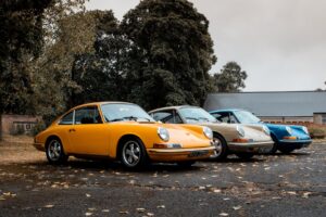 Four vintage Porsche 911 cars in yellow, beige, light gray, and blue parked in a lot with fallen leaves and trees in the background.